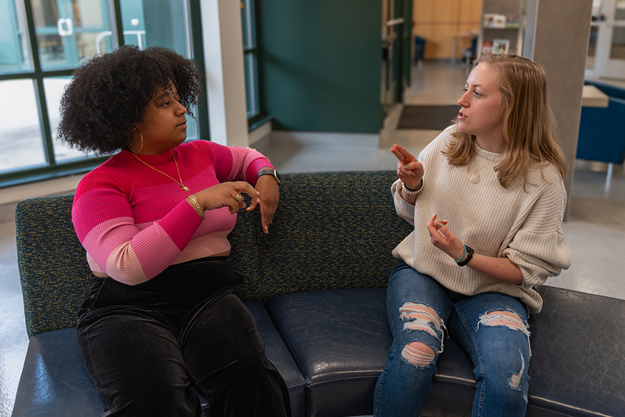 Two women sit on a dark curved sofa in a brightly lit indoor space, engaged in a conversation using American Sign Language. The woman on the left has voluminous dark curly hair and wears a vibrant pink and magenta striped sweater with gold jewelry. The woman on the right has blonde hair and wears a cream-colored ribbed sweater and distressed blue jeans. Large windows and a modern interior are visible in the soft-focus background.